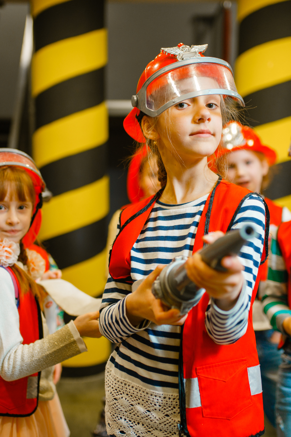 Young girl dressed in pretend firefighter equipment with toy firehouse to illustrate the importance of fire safety instruction for children.