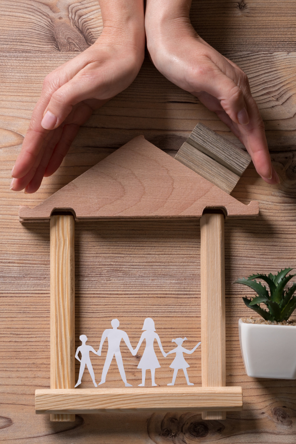 Hands forming a protective roof over a wooden house structure with a paper cutout family inside, symbolizing family safety and security.