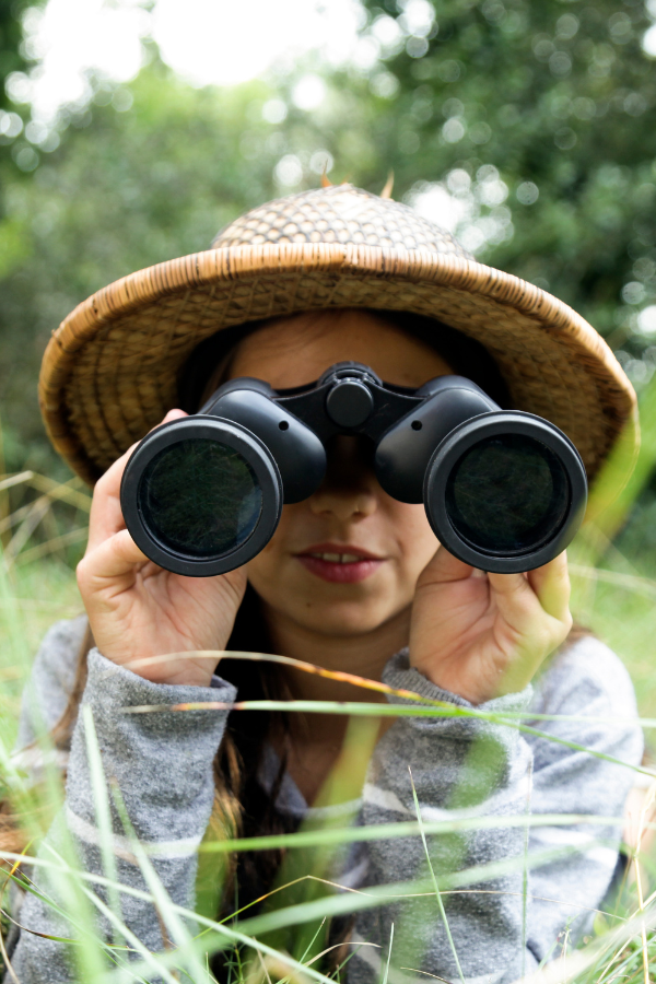 Child wearing a hat and looking through binoculars in an outdoor setting, illustrating the 'stay where you can see me' safety tip for parents and children.